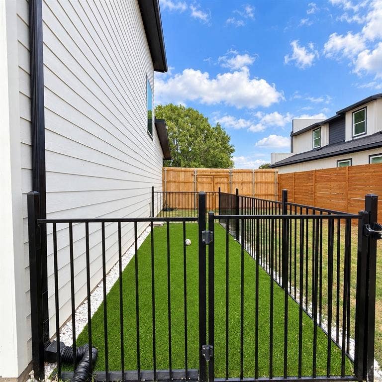 Side yard featuring synthetic turf, white stone border, black metal fencing, and wood privacy fencing
