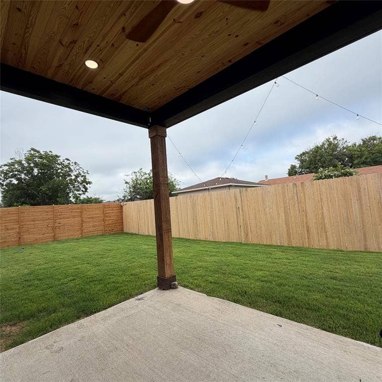 Covered patio featuring a wood-plank ceiling, recessed lighting, and a square support post, opening onto a lawn enclosed by wood fencing