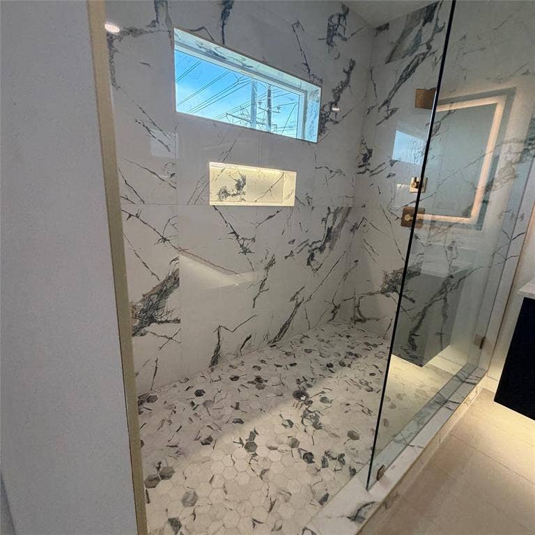 Walk-in shower featuring large-format stone-patterned tile walls, hexagonal floor tile, recessed wall LED niche, glass enclosure, and a high-set window