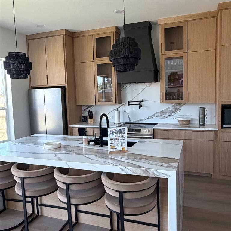 Kitchen featuring custom White Oak cabinetry, stone-patterned countertops with waterfall edge, full-height decorative backsplash, potfiller,black matte plumbing fixtures, and integrated stainless steel appliances