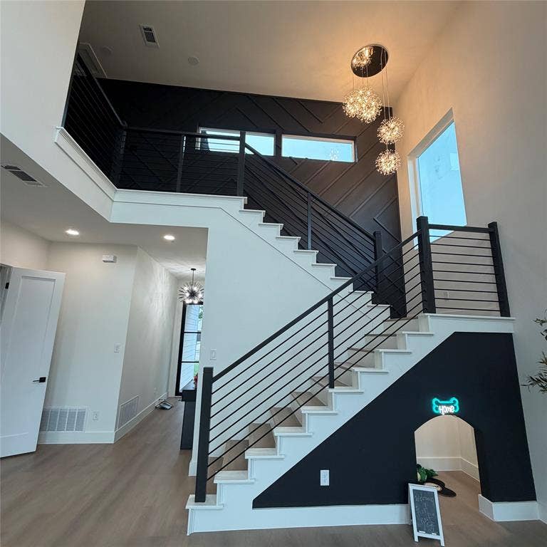 Double-height foyer featuring a staircase with horizontal metal railing, dark diagonal accent wall cladding, wood-finish flooring, and a tiered crystal chandelier