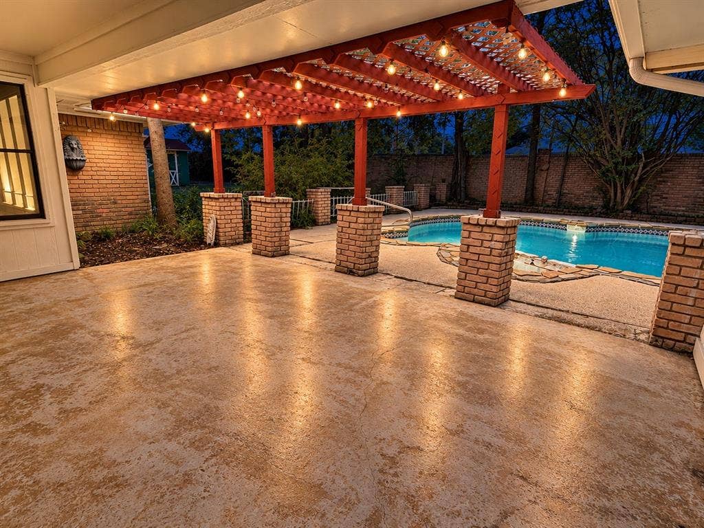 1444 Marlene Place, DeSoto, TX 75115 Covered patio featuring a red pergola with string lighting, brick support columns, textured concrete flooring, and a swimming pool with stone coping
