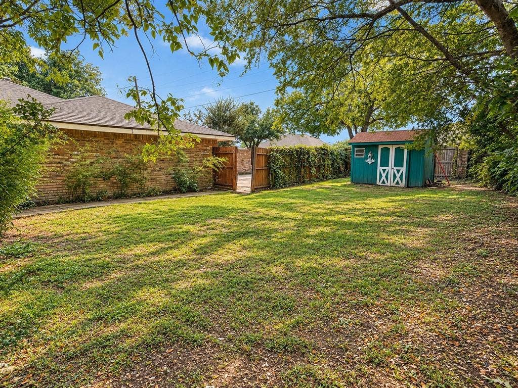 1444 Marlene Place, DeSoto, TX 75115 Expansive grassy yard with mature trees, a detached storage shed, and a brick exterior wall