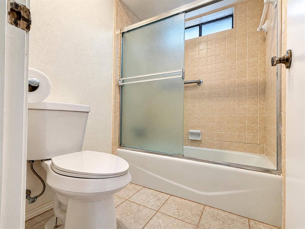 1444 Marlene Place, DeSoto, TX 75115 Bathroom featuring a white toilet, tiled flooring, tiled shower surround, and a glass-paneled tub enclosure