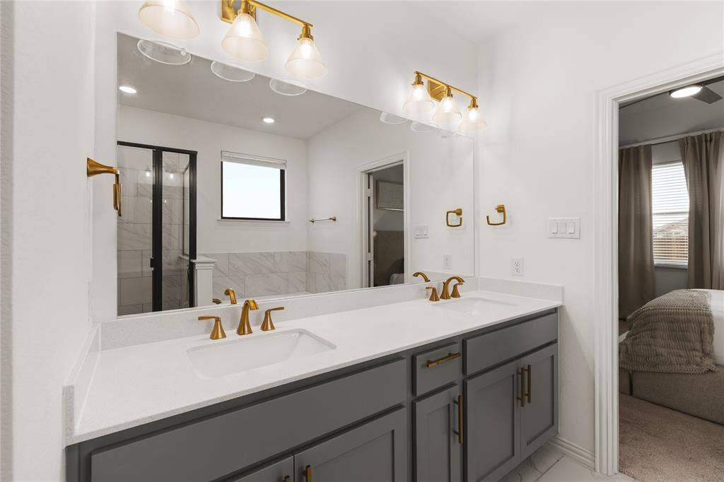 1456 Martingale Lane, Forney, TX 75126 Dual vanity with light-toned solid surface countertops, undermount sinks, brass-finish fixtures, dark cabinetry, and full-width wall mirror