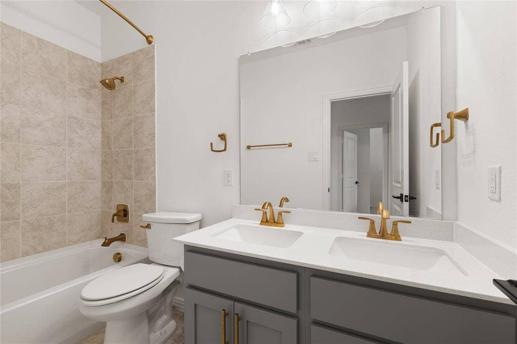 1456 Martingale Lane, Forney, TX 75126 Dual-sink vanity with quartz countertop, grey cabinetry, wall-to-wall mirror, and brass-finish hardware and fixtures, including a tiled shower-tub combination