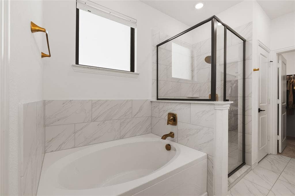 1456 Martingale Lane, Forney, TX 75126 Soaking tub with bronze-tone fixtures, glass-enclosed shower with a matte black frame, and large-format stone-patterned tile wall surrounds