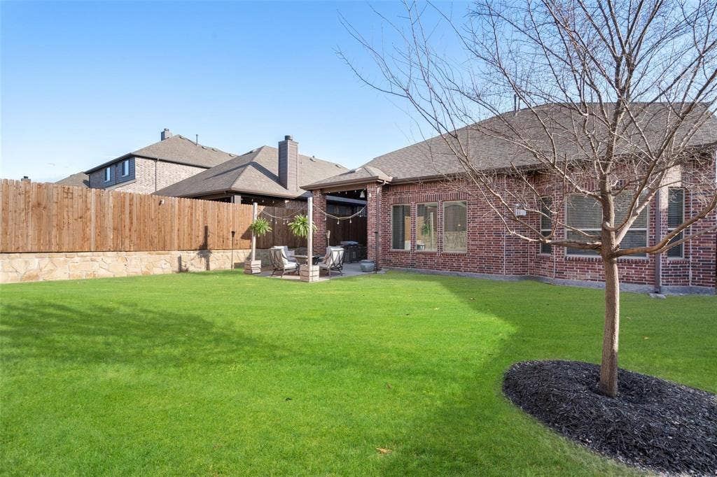 1017 Hoyt Drive, McKinney, TX 75071 Expansive backyard featuring a manicured lawn, brick exterior, covered patio, and privacy fencing with stone foundation