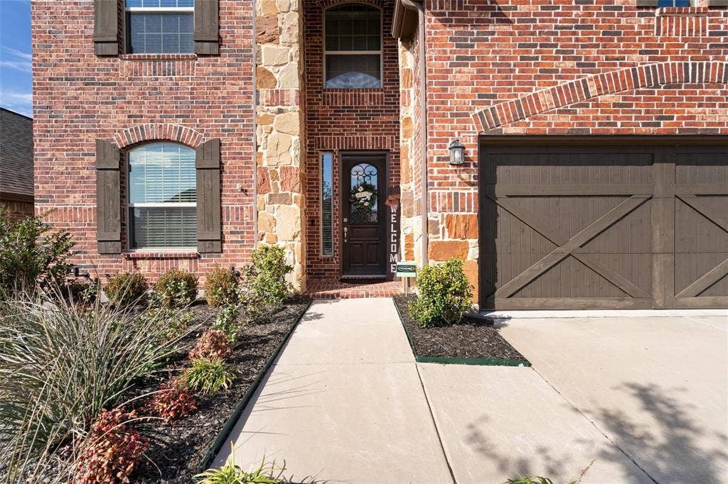 1017 Hoyt Drive, McKinney, TX 75071 Brick and stone exterior featuring an arched entryway, decorative wooden garage door, and established landscaping