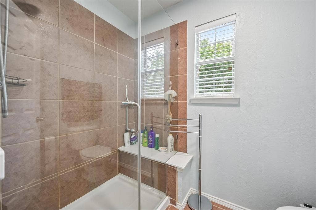 2209 Stonepark Place, McKinney, TX 75071 Glass-enclosed shower with a built-in bench, brown tile surround, and dual windows with blinds