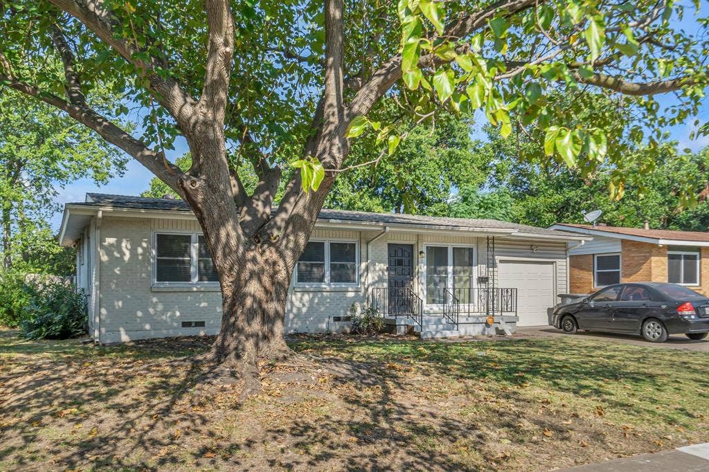 9621 Rustown Drive, Dallas, TX 75228 Exterior featuring a large mature tree, light-colored stucco siding, a covered front entry with railing, and an attached garage with a corrugated door