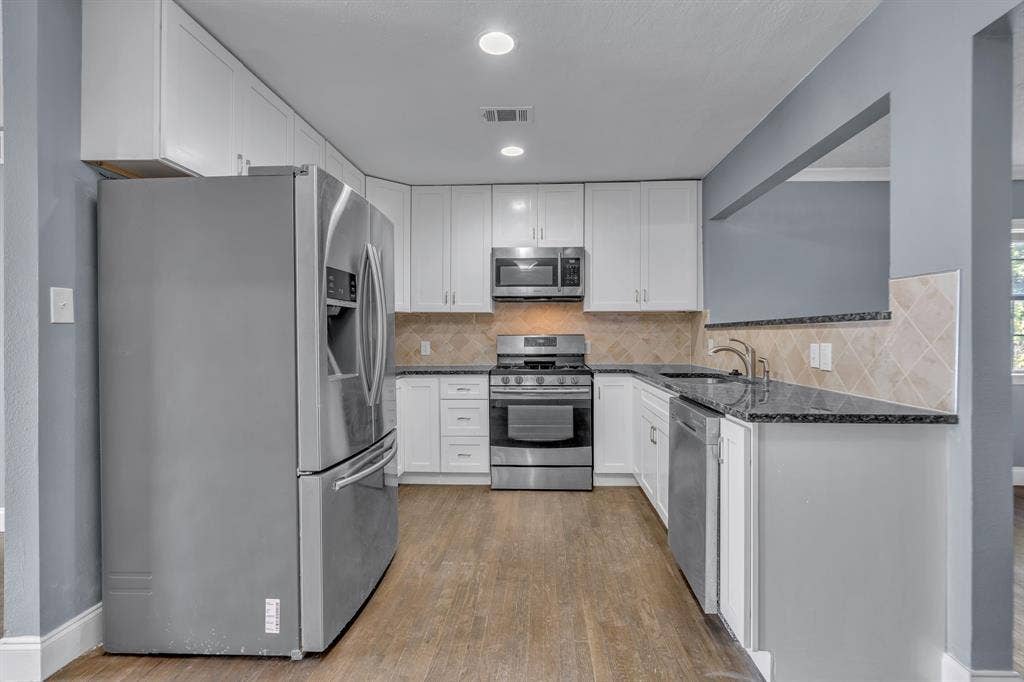 Kitchen featuring stainless steel appliances, dark countertops, white cabinetry, tile backsplash, and wood-finish flooring