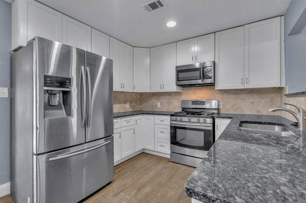 Kitchen featuring white shaker cabinetry, stainless steel appliances, dark stone countertops, tile backsplash, and wood-finish flooring