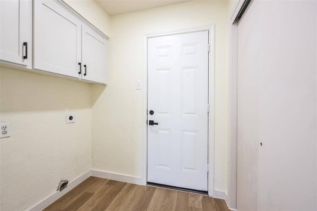 Wood-finish flooring with white baseboards, white paneled door with black hardware, and white upper cabinetry with black pulls