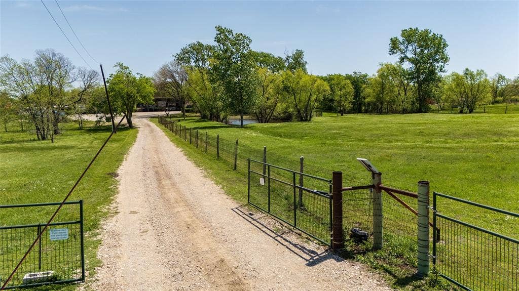 Gravel driveway entry featuring a metal perimeter fence, expansive open acreage, and mature trees
