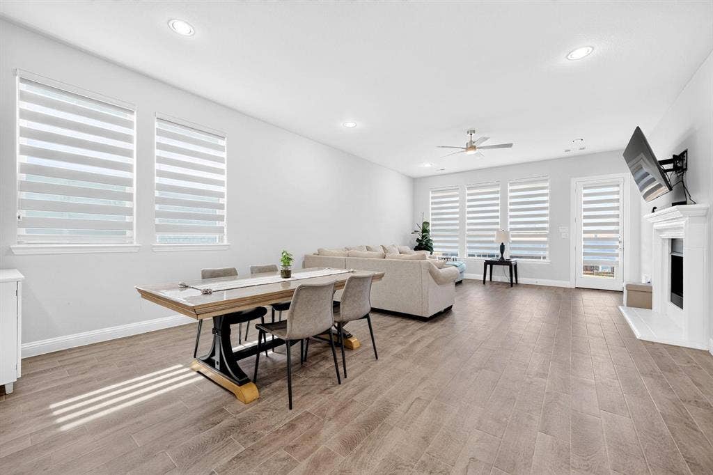 Open-concept space featuring wood-finish flooring, recessed lighting, a ceiling fan, tiered window shades, and a fireplace with a white mantel
