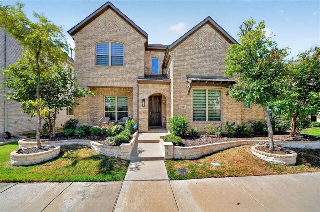 Two-story brick exterior featuring a covered entryway, decorative stone-walled garden beds, dual gabled rooflines, and multi-pane windows