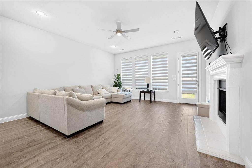 Living area featuring wood-finish flooring, recessed lighting, ceiling fan, triple-pane window configuration with integrated blinds, and a white hearth fireplace