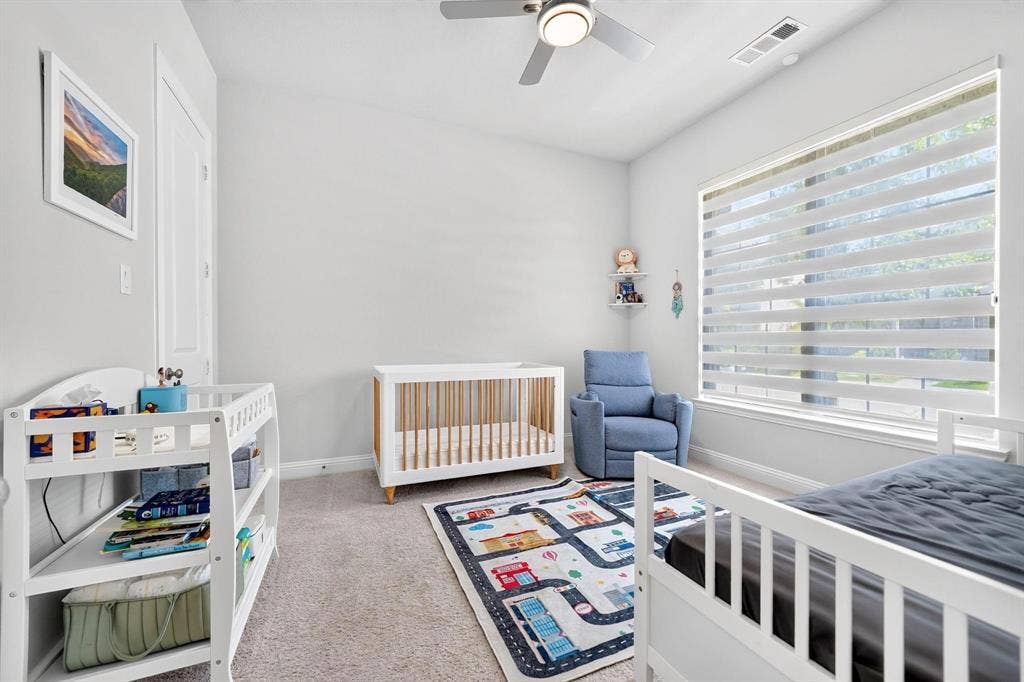 Neutral-toned interior featuring plush carpeting, a multi-blade ceiling fan with integrated lighting, and a large window fitted with horizontal light-filtering shades