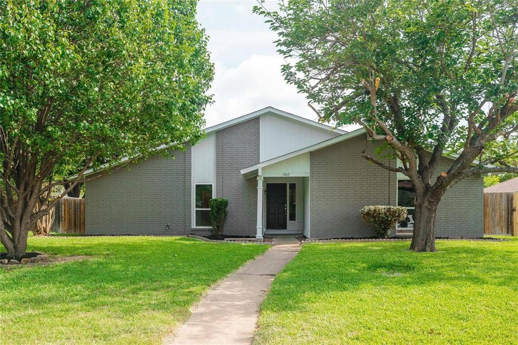1903 Arvada Drive, Richardson, TX 75081 Single-story residence featuring a painted brick exterior, gabled roofline, front-facing vertical siding accents, and mature shade trees