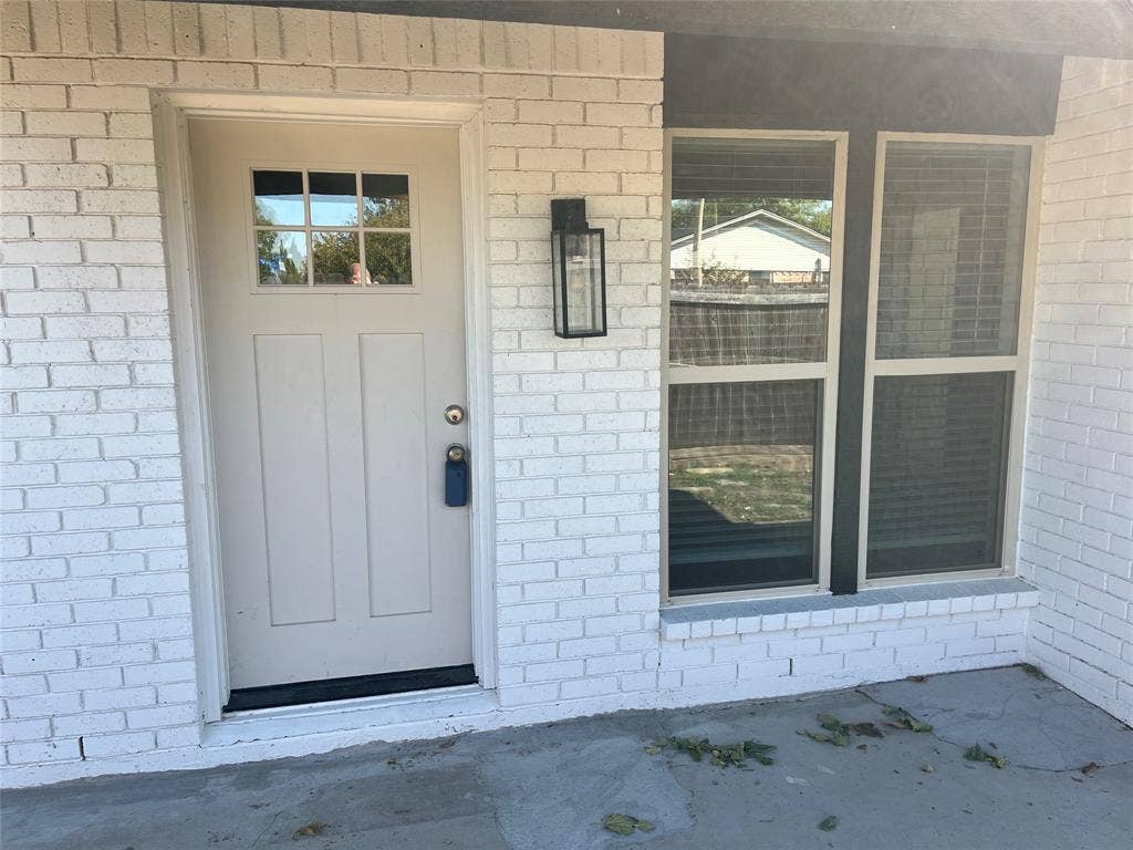 600 Royal Crest Drive, Richardson, TX 75081 White brick exterior featuring a single-panel front door with a six-pane window insert, paired with dual windows and a rectangular matte-black wall-mounted lantern