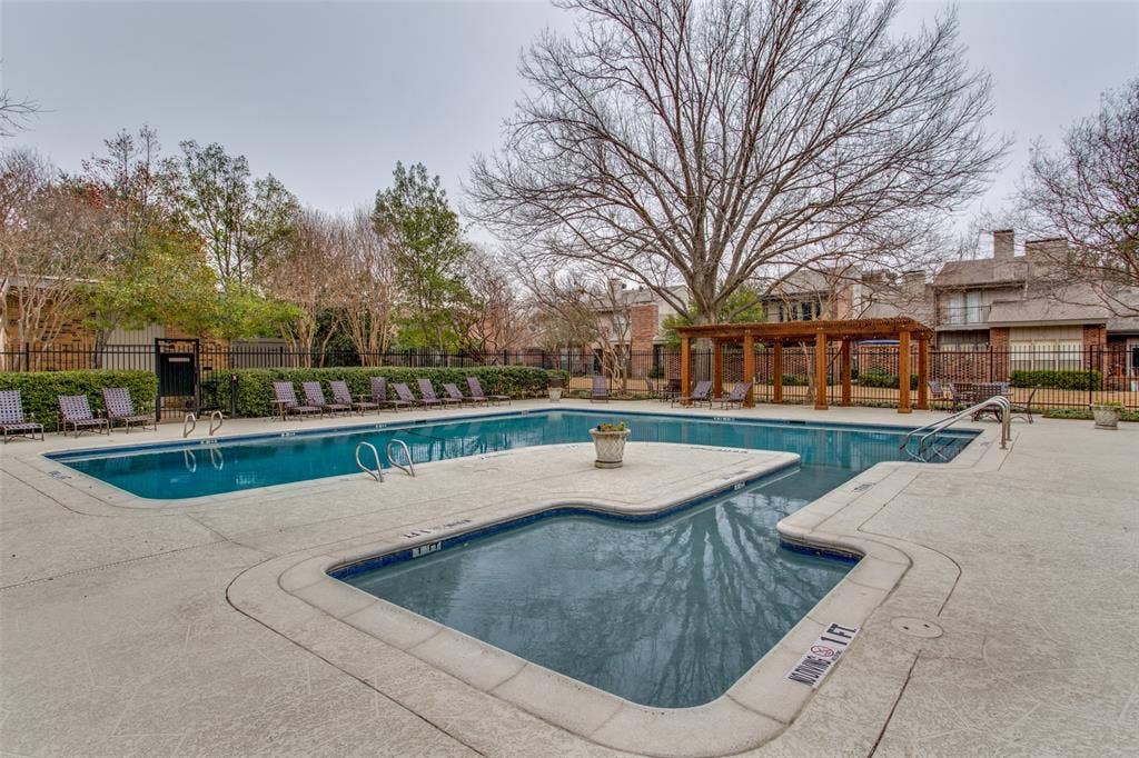 In-ground swimming pool with shallow wading area, surrounding concrete decking, wood-frame pergola, and perimeter metal fencing