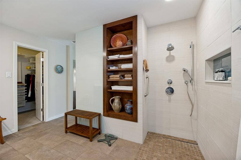 Walk-in shower with large-format wall tile, mosaic tile floor, and chrome fixtures featuring a rainfall showerhead and handheld sprayer, complemented by built-in wood shelving and an integrated tile shower niche