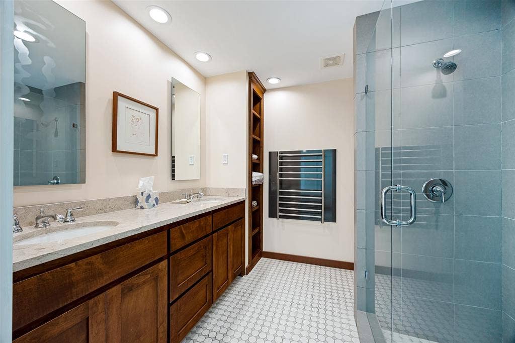 Double vanity with wood-finish cabinetry, stone countertop, recessed lighting, built-in floor-to-ceiling shelving, and a glass-enclosed shower featuring tile walls