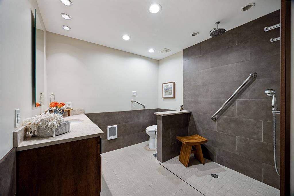 Bathroom featuring dark wood-finish cabinetry, a stone-surface countertop, recessed lighting, and a walk-in shower with large-format tile surround, integrated grab bars, and a wall-mounted shower fixture