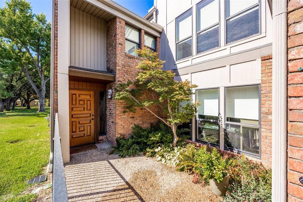 Exterior entrance featuring a decorative wood front door, multi-tonal brick masonry, vertical siding, large multi-pane windows, and a landscaped front garden bed