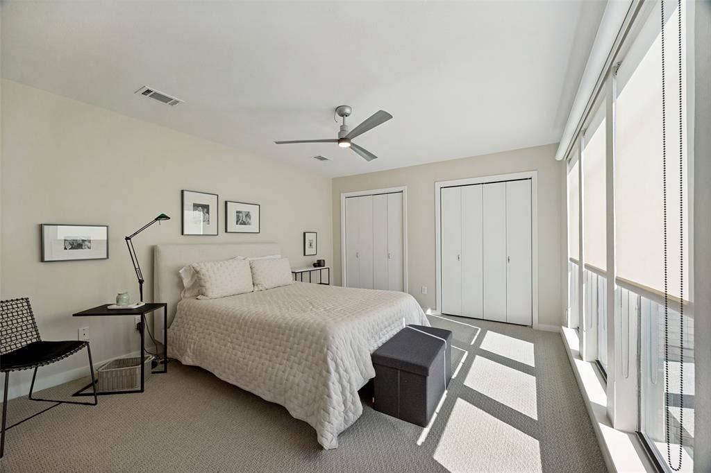 Room featuring neutral wall-to-wall carpeting, floor-to-ceiling windows with roller shades, a ceiling fan with integrated lighting, and dual closets with bifold doors