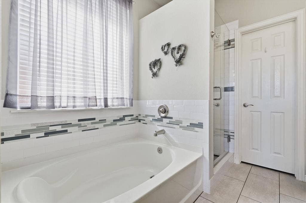 A soaking tub beneath a large window offers a relaxing spot, with decorative tile continuing into the separate shower.