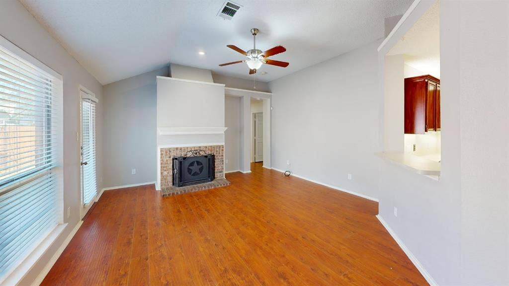 5210 Welbeck Court, Garland, TX 75043 Vaulted ceiling living area featuring wood-finish flooring, a brick fireplace with a mantle, a ceiling fan, and a pass-through window to the kitchen