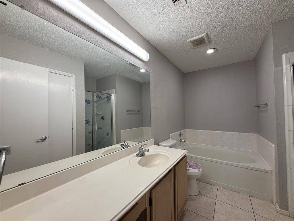 5210 Welbeck Court, Garland, TX 75043 Full-width wall mirror, wood-finish vanity cabinet with a neutral integrated countertop and sink, soaking tub with a tiled surround, and ceramic tile flooring