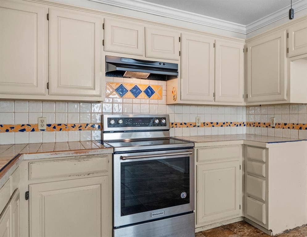 Kitchen featuring painted cabinetry, a stainless steel range, a mounted range hood, tiled countertops with decorative trim, and crown molding