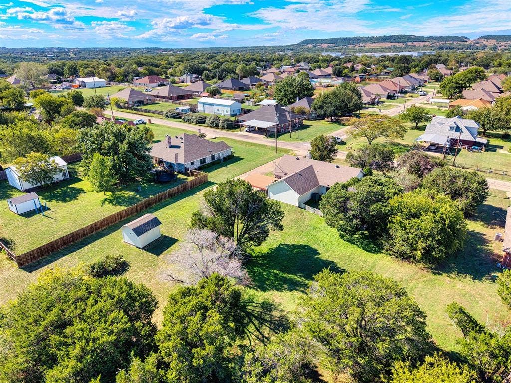 Aerial perspective of a multi-structure property featuring a expansive grassy lot, mature trees, a detached storage building, and a wood privacy fence