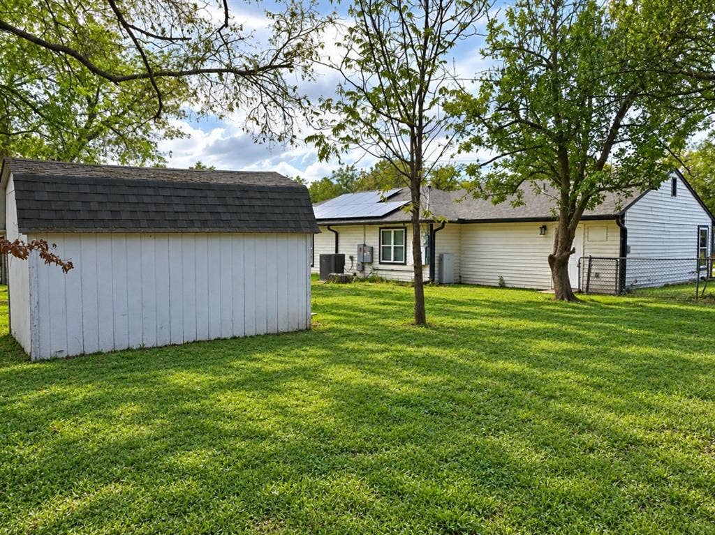 709 SW Dallas Street, Grand Prairie, TX 75051 Spacious backyard featuring a detached storage structure, mature trees, chain-link fencing, and a residence equipped with roof-mounted solar panels