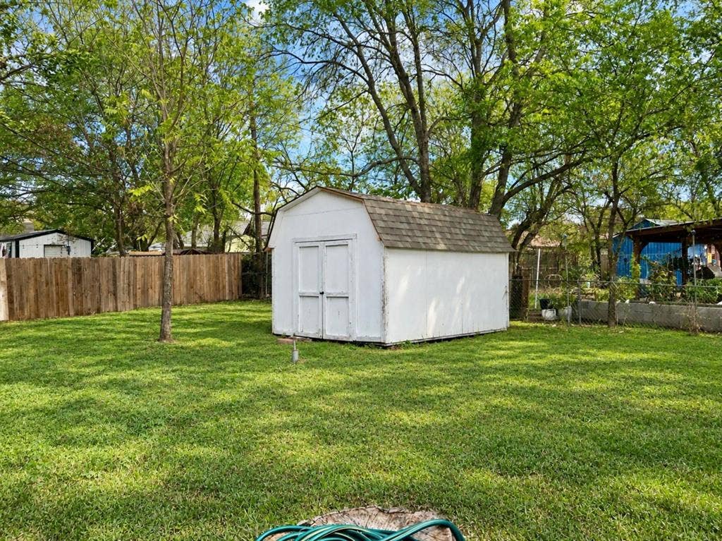 709 SW Dallas Street, Grand Prairie, TX 75051 Backyard area featuring a white, gambrel-style storage structure with asphalt shingle roofing, enclosed by a wood privacy fence and established trees