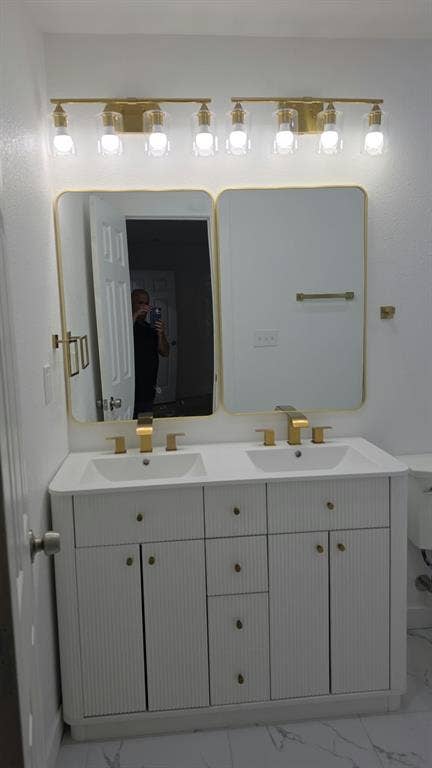 3426 Edgewood Street, Dallas, TX 75215 Dual-sink vanity featuring a white fluted cabinet, stone-finish countertop, brass hardware, coordinating dual gold-framed mirrors, and a multi-light brass fixture