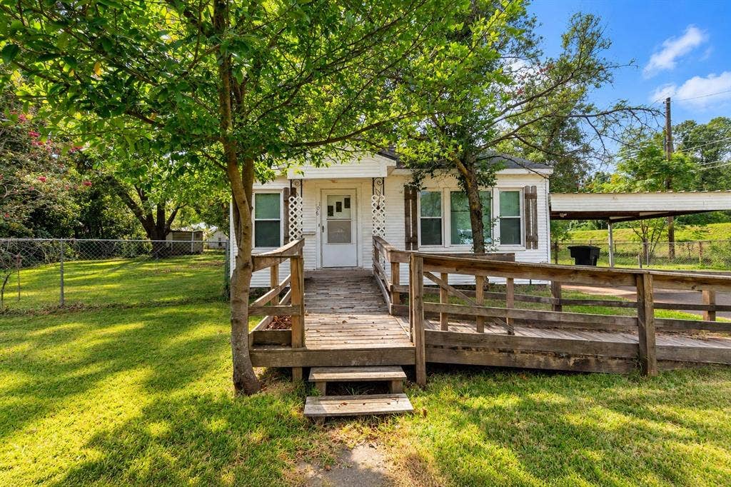 White siding exterior featuring a wooden ramp, attached carport, chain-link perimeter fencing, and mature trees