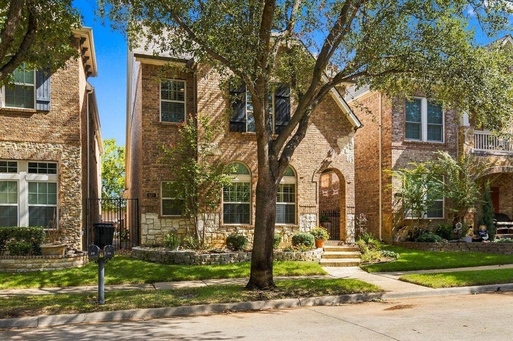 Brick and stone veneer exterior featuring arched entryway, decorative window shutters, and manicured landscaping with mature trees