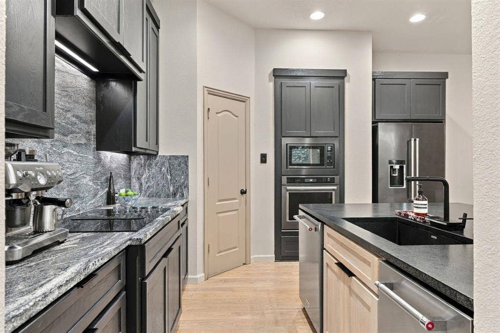 Kitchen featuring charcoal cabinetry, natural stone countertops with matching backsplash, built-in stainless steel double oven, and wood-finish flooring