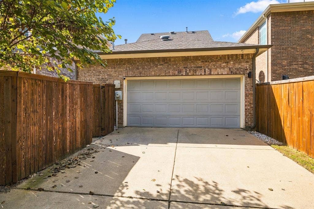 Two-car garage with brick exterior, composite shingle roof, paneled door, and concrete driveway enclosed by wood fencing