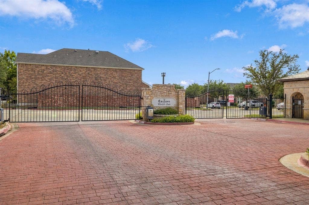 Gated entrance featuring red brick paver driveway, ornate wrought-iron security gate, stone monument signage, and perimeter brick wall