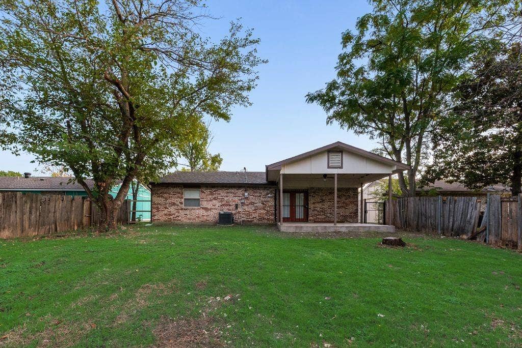 Rear exterior featuring a brick facade, covered patio, grass lawn, mature trees, and wood fencing