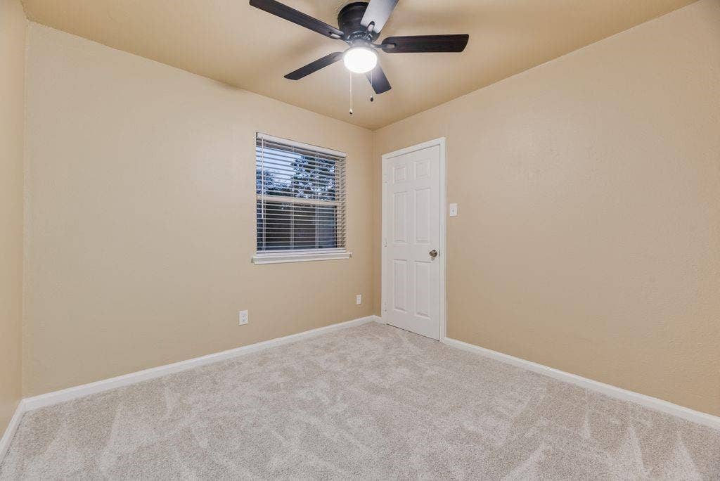Neutral wall paint, plush carpeting, multi-pane window with horizontal blinds, six-panel interior door, and integrated ceiling fan with light fixture