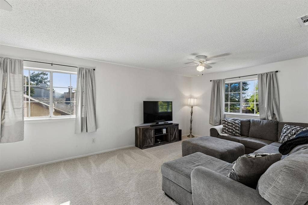 Neutral-toned interior space featuring light-colored wall-to-wall carpeting, dual windows with multi-pane grids, and a ceiling-mounted light fixture with a multi-blade fan