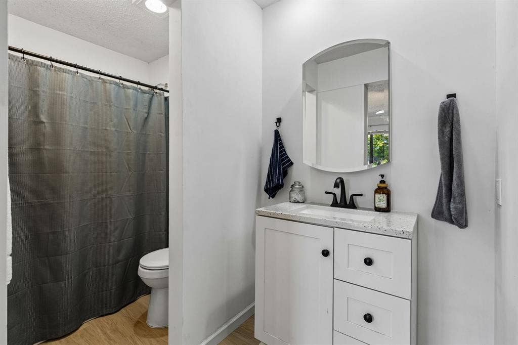 Bathroom featuring a white vanity cabinet with stone-look countertop, rectangular under-mount sink, matte black hardware, arched wall-mounted mirror, and wood-finish flooring