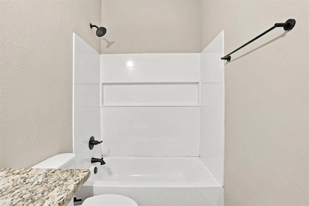 1116 S 6th Avenue, Denison, TX 75021 Bathtub and shower combination featuring white surround paneling with integrated recessed shelving, matte black fixtures, and a stone-finish countertop