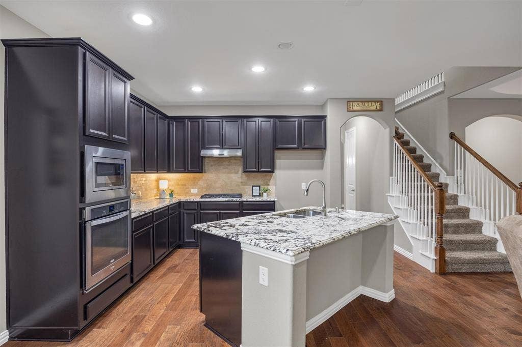 3014 Aurora Mist Drive, Little Elm, TX 75068 Kitchen featuring 42-inch dark cabinetry, granite countertops, stainless steel built-in oven and microwave, center island with a dual-basin sink, and wood-finish flooring